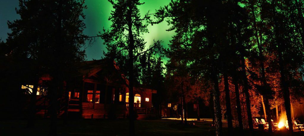 Outside image of the lodge with a campfire to the right and the Canadian Northern Lights in the background to the left