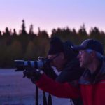 Two people, one person looking into a camera lens and the other coaching what to look for or do