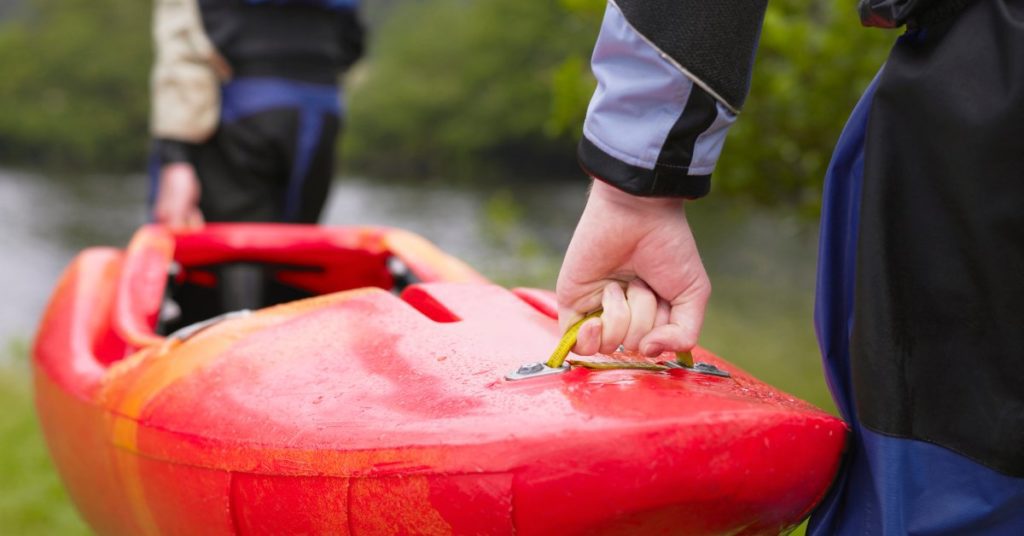 Two people carrying a large red kayak outdoors toward a nearby river surrounded by many massive green trees.