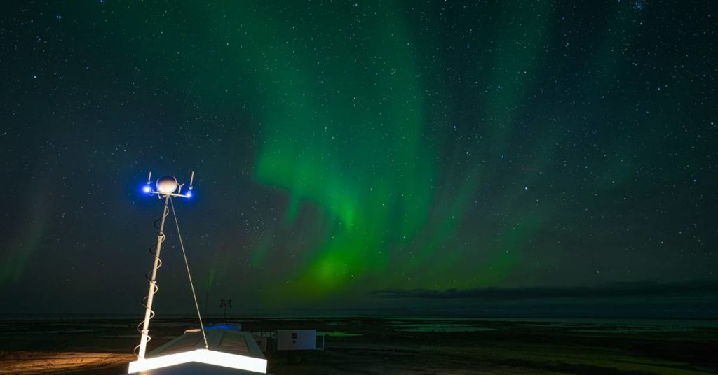 The northern lights shining bright green in the night sky in Manitoba. Several buildings sit idly below.